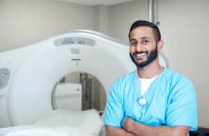 Young adult radiologist smiles while standing next to CT machine